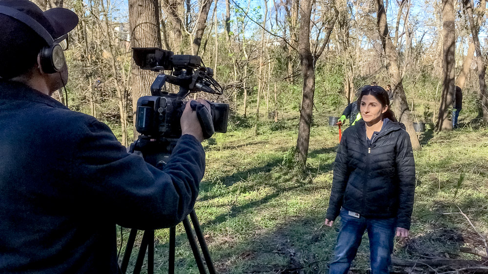 Kelli Ondracek, '11 MS A person being interviewed on camera, standing in the middle of a riparian habitat with trees and vegetation surrounding them.