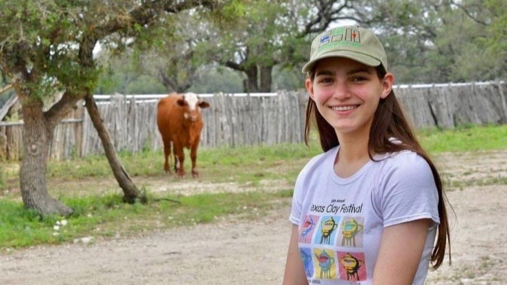 An individual wearing a cap. In the background, a solitary cow stands in a field surrounded by trees and a fence line.