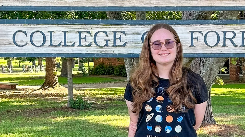 Person in a black t-shirt with colorful planet designs standing in front of a wooden “College of Forestry” sign.