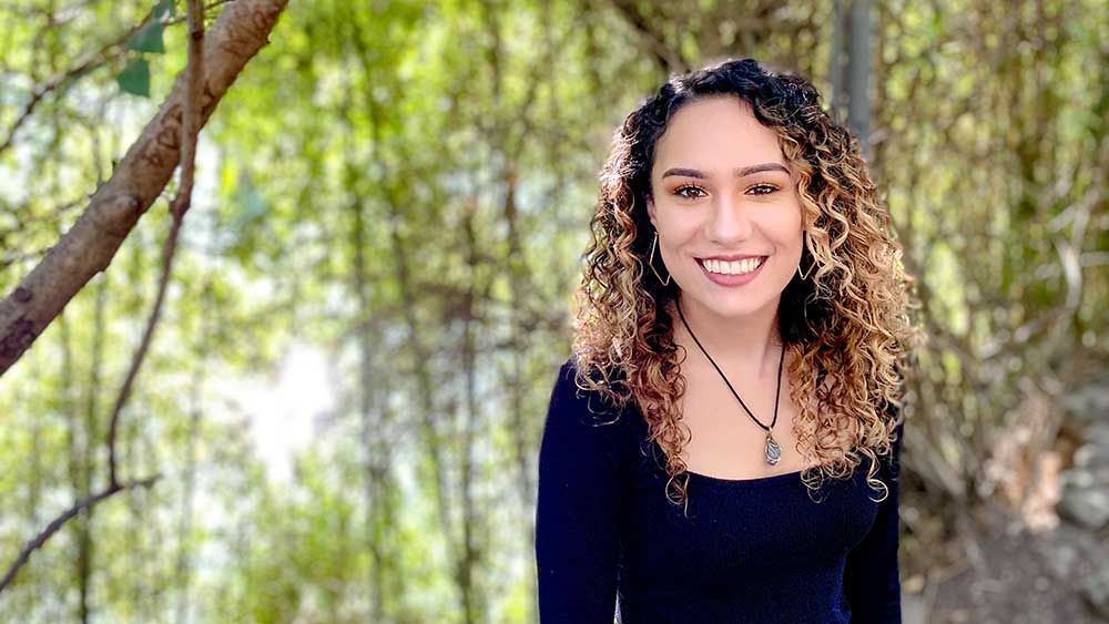 Tayler McKenzie Tayler is standing in front of a vibrant green forest. She is smiling at the camera.