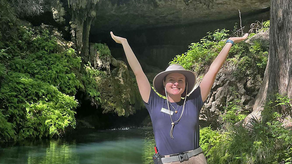 Loralei Miller An individual standing with arms raised in a carefree gesture in front of a serene body of water, with greenery and an overhang in the background.