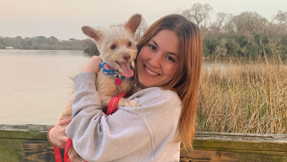Tatum Morgan An individual, holding a dog, stand in front of a body of water, surrounded by tall grasses.