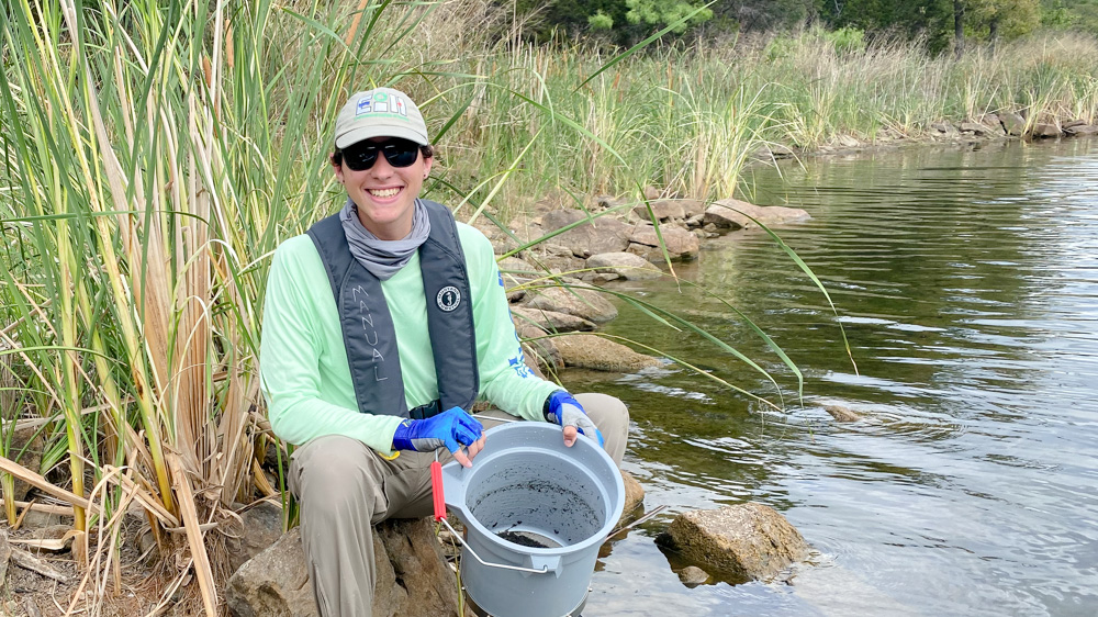Chris is sitting on the bank of a stream holding a sieve bucket and smiling at the camera. Chris Pryor