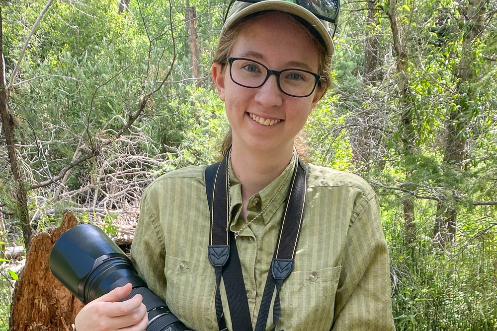 Person wearing glasses, a cap with sunglasses on top, and a green shirt, smiling while holding a camera with a large telephoto lens in a forest with dense green foliage and tree branches.