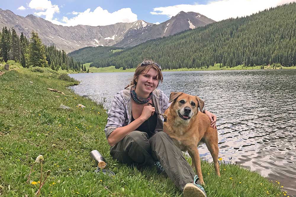 Sarah Schrock Sarah is sitting on a grassy bank of a lake with her dog with an evergreen forest and mountains in the background.