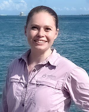 Melanie Rogers Melanie is standing in front of a large body of water on a sunny day.