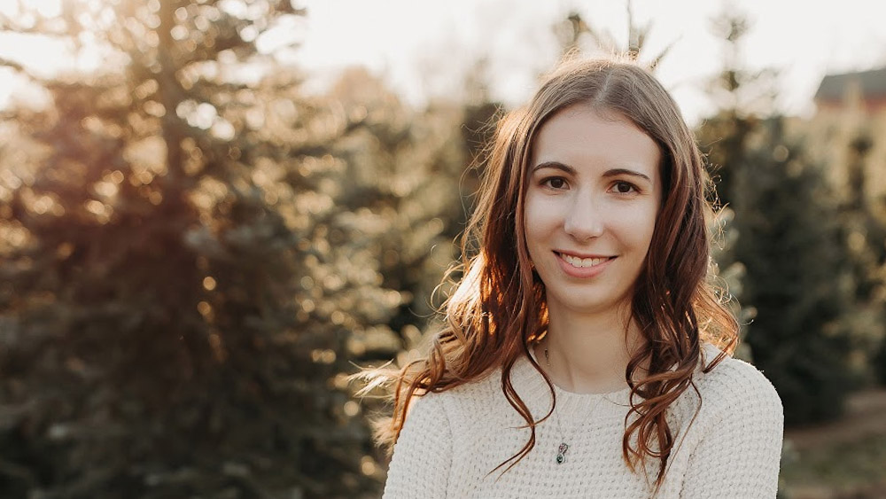 Hanah Martin Person with long wavy hair wearing a light sweater, smiling outdoors with evergreen trees in the background.