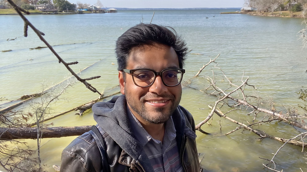 A person in glasses and a jacket smiling by calm water, with fallen branches in the water and a tree-lined shore in the distance.