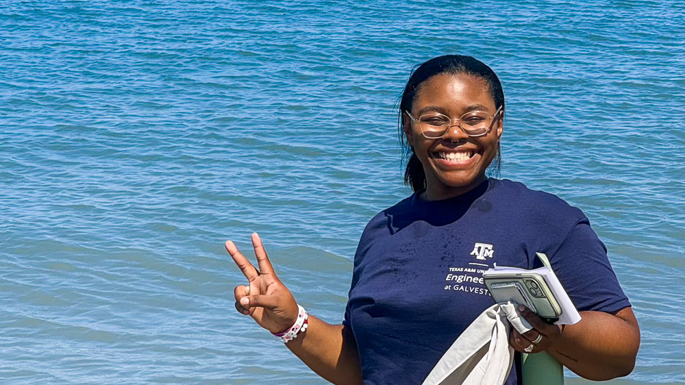 Person standing by calm blue water, holding a phone and a white towel in one hand while making a peace sign with the other.