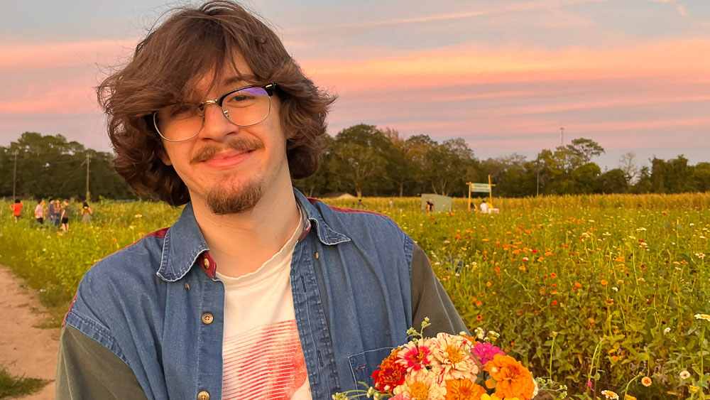 Luke Hammock Luke happily stands in a field of flowers, holding a colorful bouquet. He smiles at the camera against a stunning blue and pink sky.