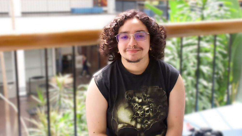A seated person with shoulder‑length curly hair and glasses sits indoors near a wooden railing, with green plants and a blurred building interior in the background.