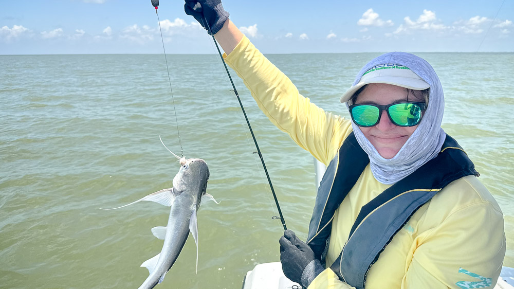 A person on a boat, wearing a long-sleeve yellow shirt, sunglasses, gloves, and a life jacket, holds up a fishing line with a small catfish over green coastal water.