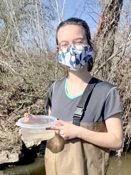 Catherine Bradley Catherine is masked up and holding a container filled with water and a few small fish. In the background is a stream bank covered in bare stems.