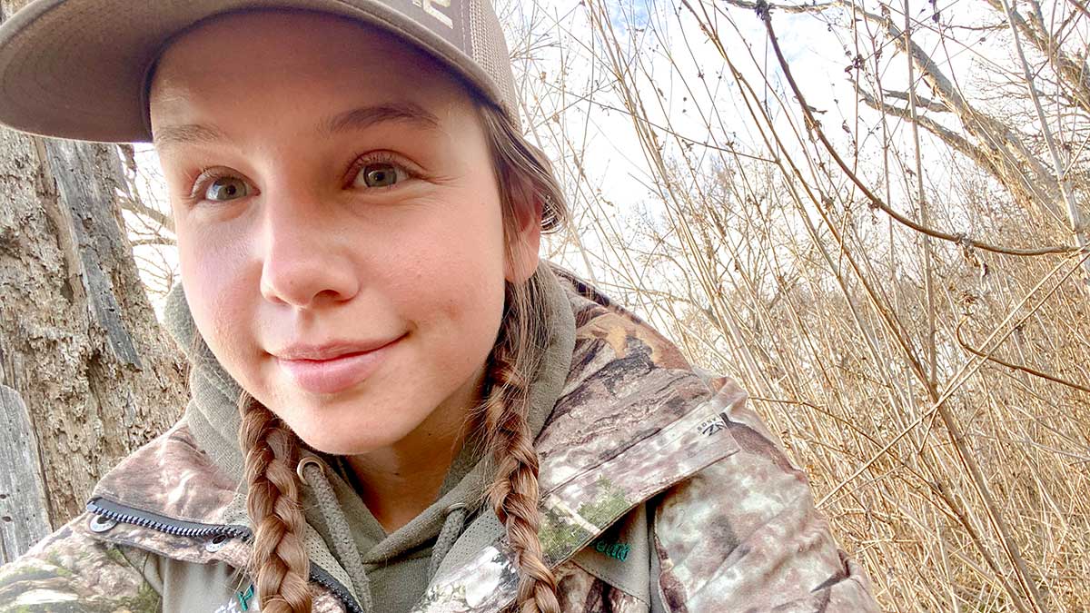 Joanna Fuess Joanna is outside in a wooded environment. She is wearing camouflage field gear.