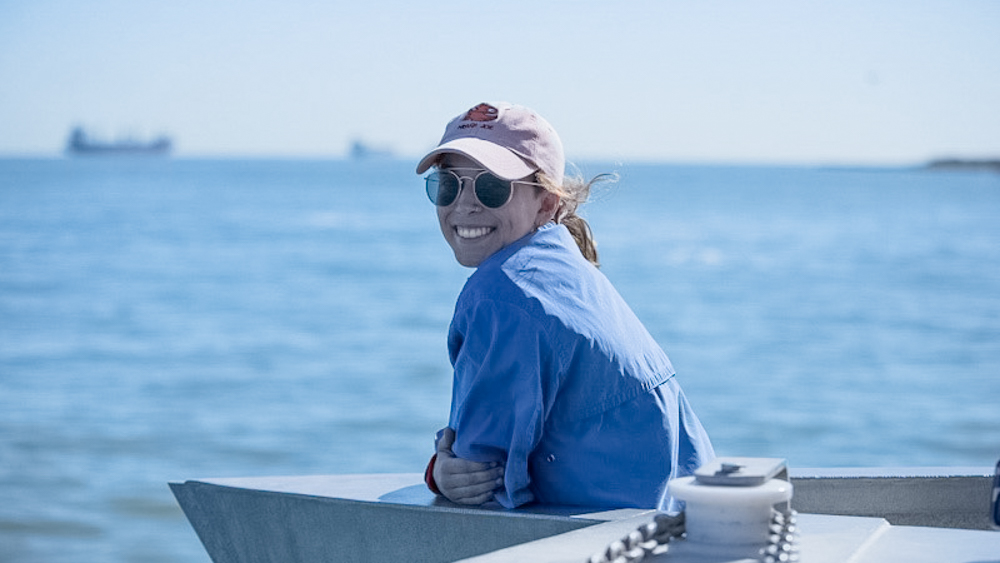 Taryn Hood Taryn is on a ship and smiling at the camera with the ocean and distant ships in the background.