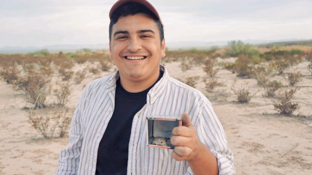 Nick is in a desert habitat holding a small rodent trap in his left hand. He is smiling at the camera. Nick Hughes