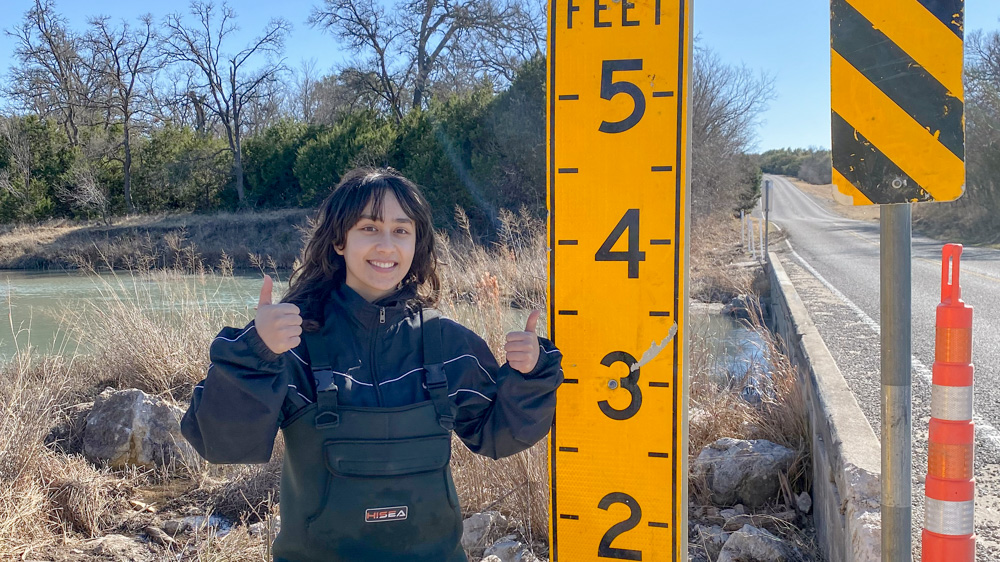 Adriana Puzon Adriana is giving two thumbs up from where she stands next to a flood gauge.