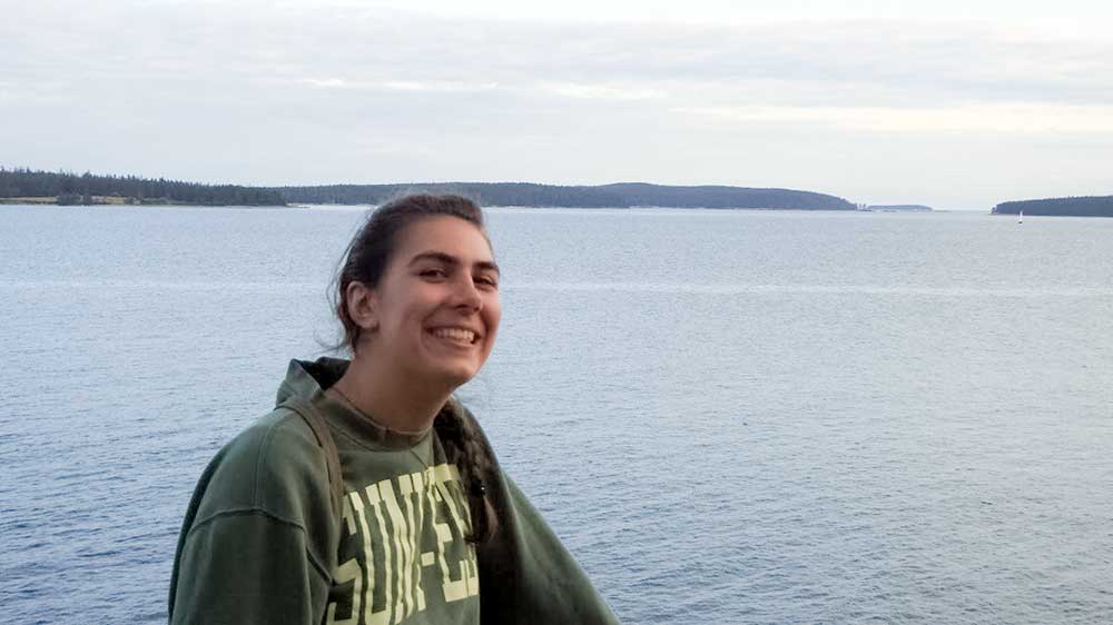 Haley Welshoff Haley is smiling into the camera. Behind her is a large body of water with tree-covered hills in the horizon.
